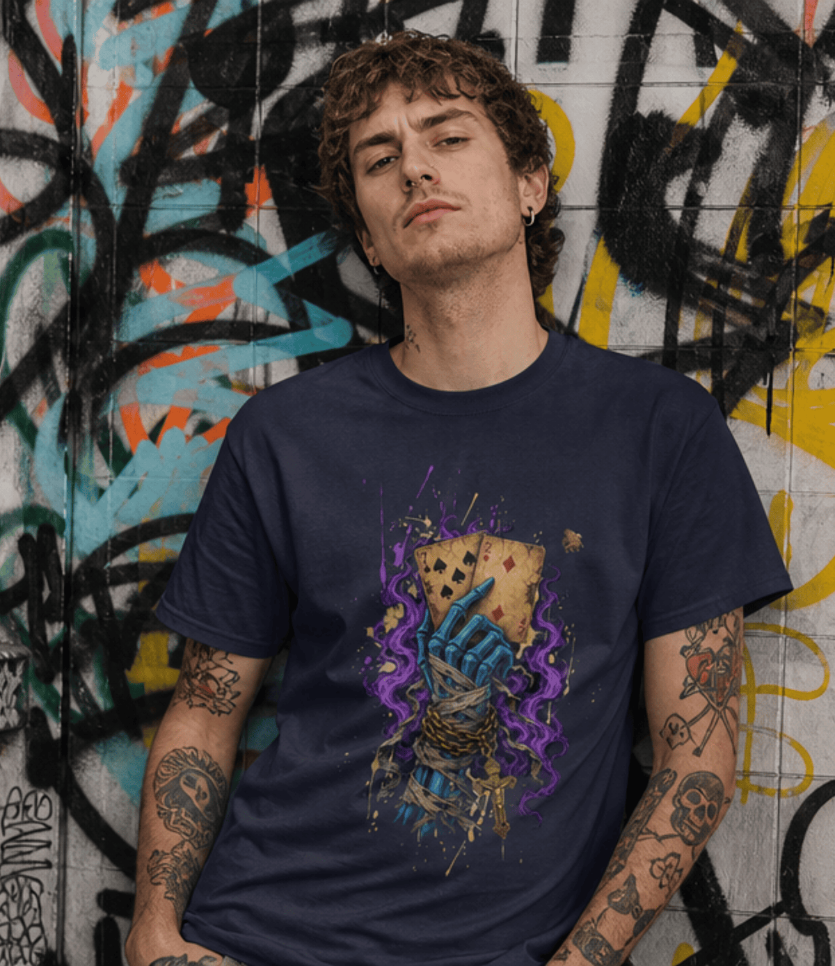 A man with tattoos leans against a graffiti-covered wall wearing a navy blue t-shirt with a graphic of a skeletal hand gripping playing cards, surrounded by purple and gold paint splatters.