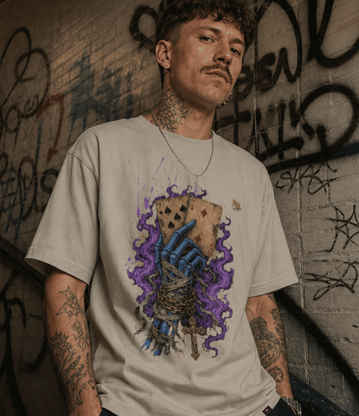 A man with tattoos and a mustache wearing a light beige graphic t-shirt, standing against a graffiti-covered brick wall. The t-shirt features a graphic of a blue skeletal hand holding a playing card, surrounded by purple smoke.