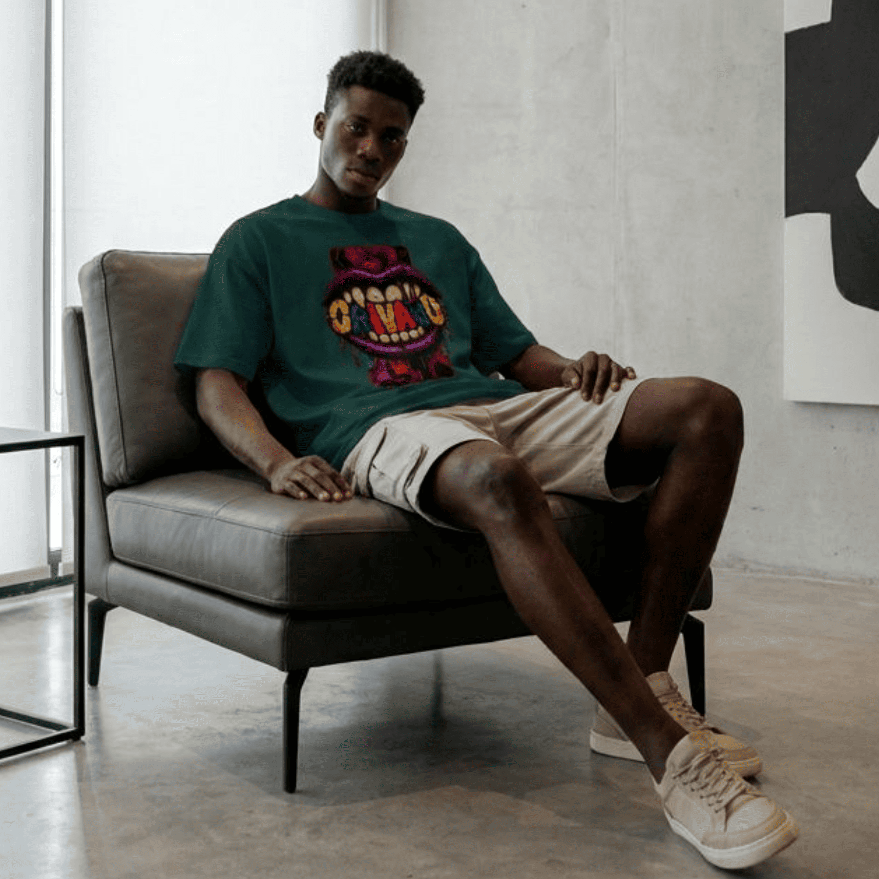 A man sits in a grey leather armchair wearing a dark green graphic t-shirt with a colourful crocodile design, beige shorts, and white sneakers in a modern, minimalist room with concrete floors and walls.