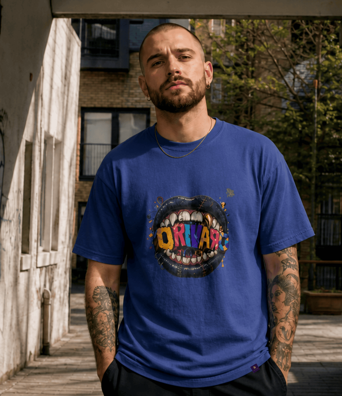 A man wearing a royal blue graphic t-shirt with a colourful 'ORIVARO' design inside a circular mouth graphic, standing outdoors in a sunny alleyway.