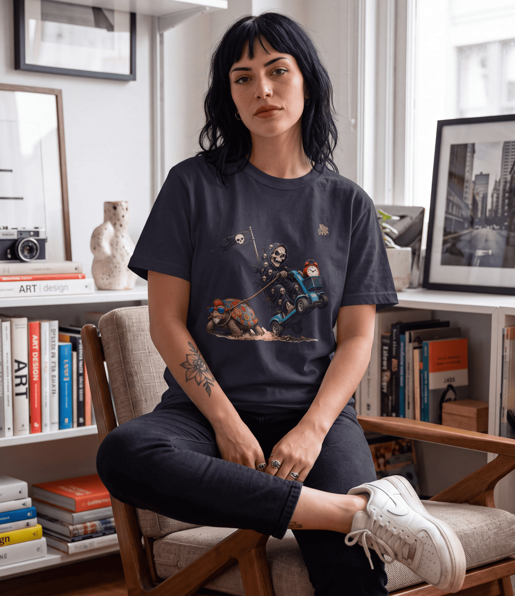 A woman sits in a wooden armchair in a bright, modern room, wearing a dark charcoal graphic t-shirt with a detailed illustration of a knight in armour on a blue vehicle. She is also wearing black pants and white sneakers.
