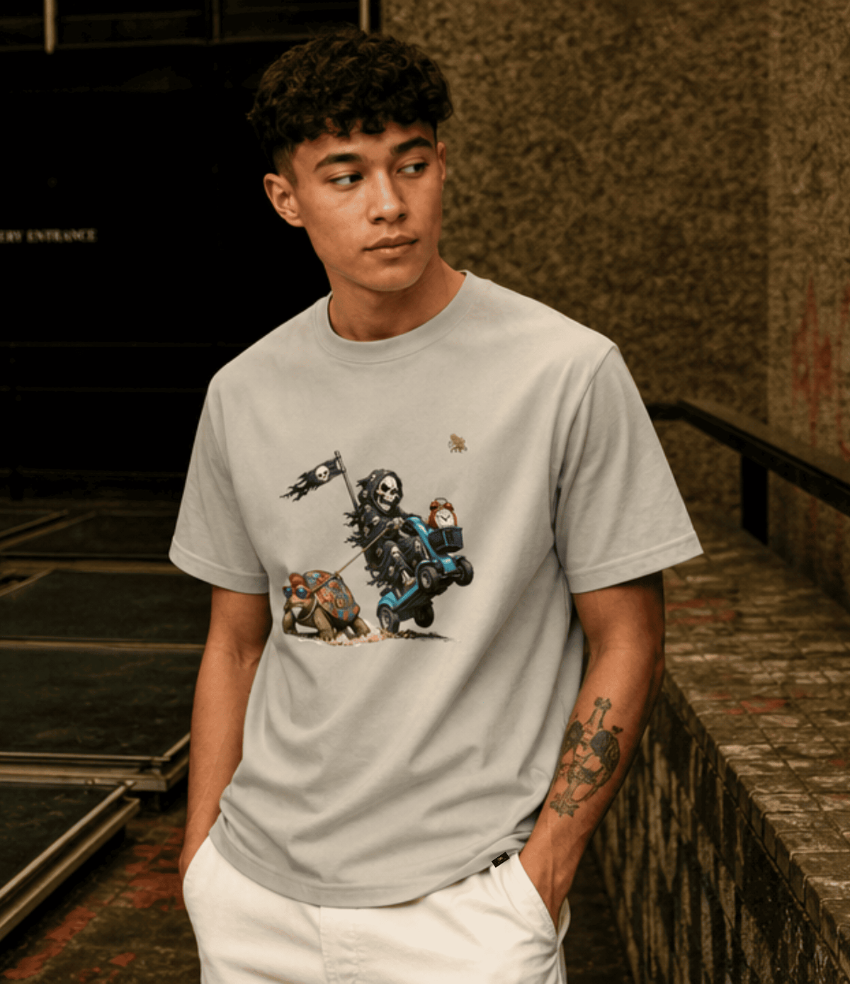 A man wearing a light grey crewneck t-shirt with a detailed graphic print of a skeleton knight on a blue cart, paired with white pants, standing in a dimly lit urban alleyway.