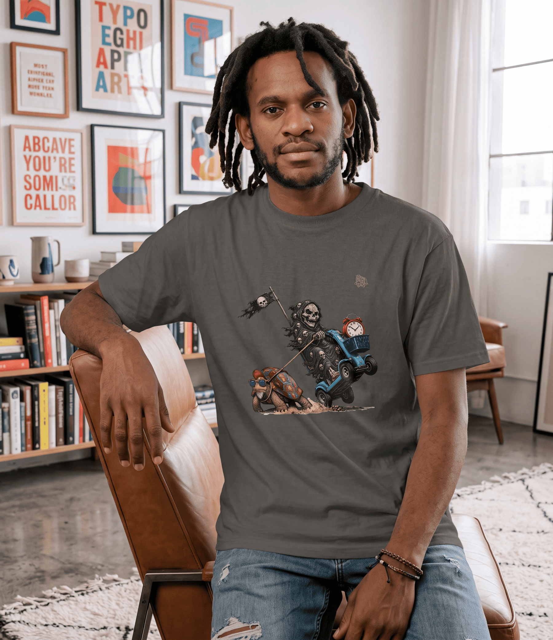 A man with dreadlocks sits in a brown leather chair wearing a dark grey graphic t-shirt with a cartoon skeleton character. The background shows a cozy, modern living room with framed art on the wall, a bookshelf, and a window.