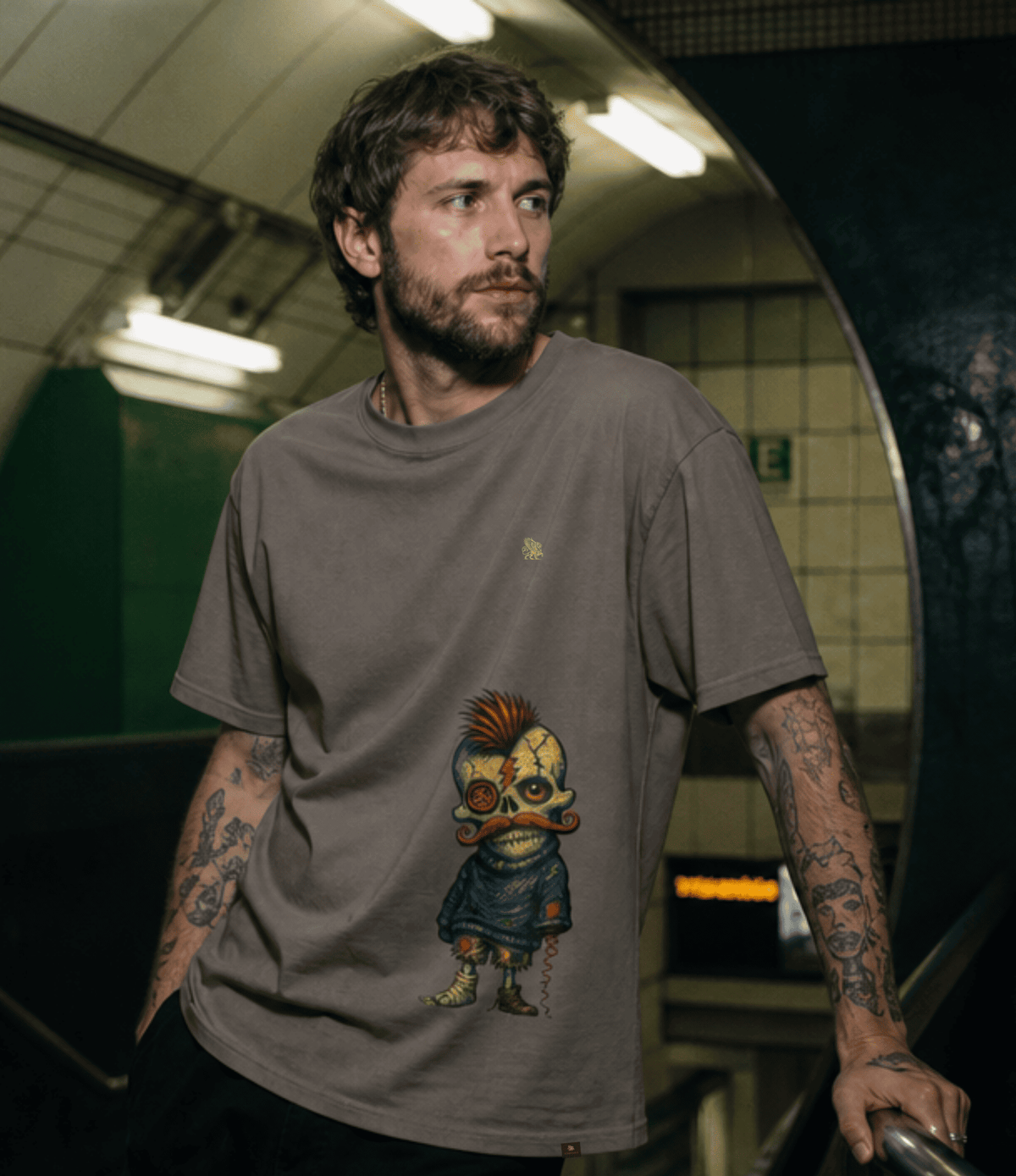 A man with tattoos wearing a brownish-grey graphic t-shirt featuring a cartoon character, standing in a subway station.