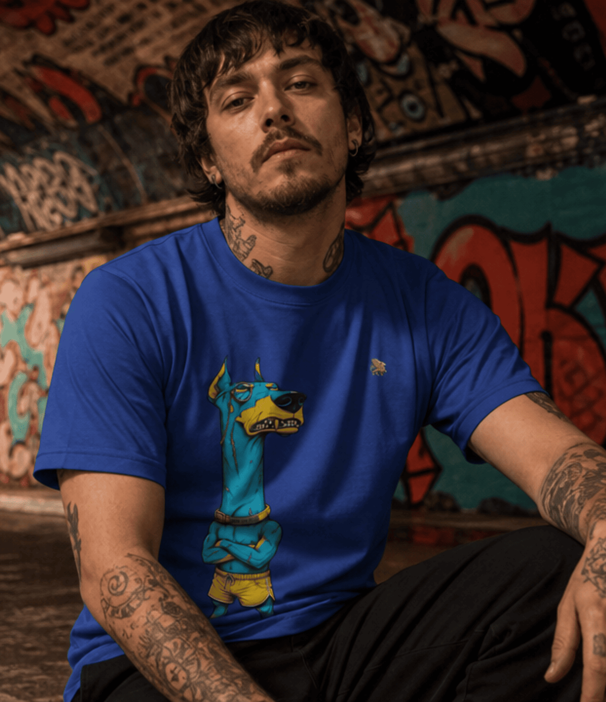 A man with tattoos wearing a royal blue t-shirt featuring a cartoon graphic of a blue and yellow dog, posed in front of a graffiti-covered wall.