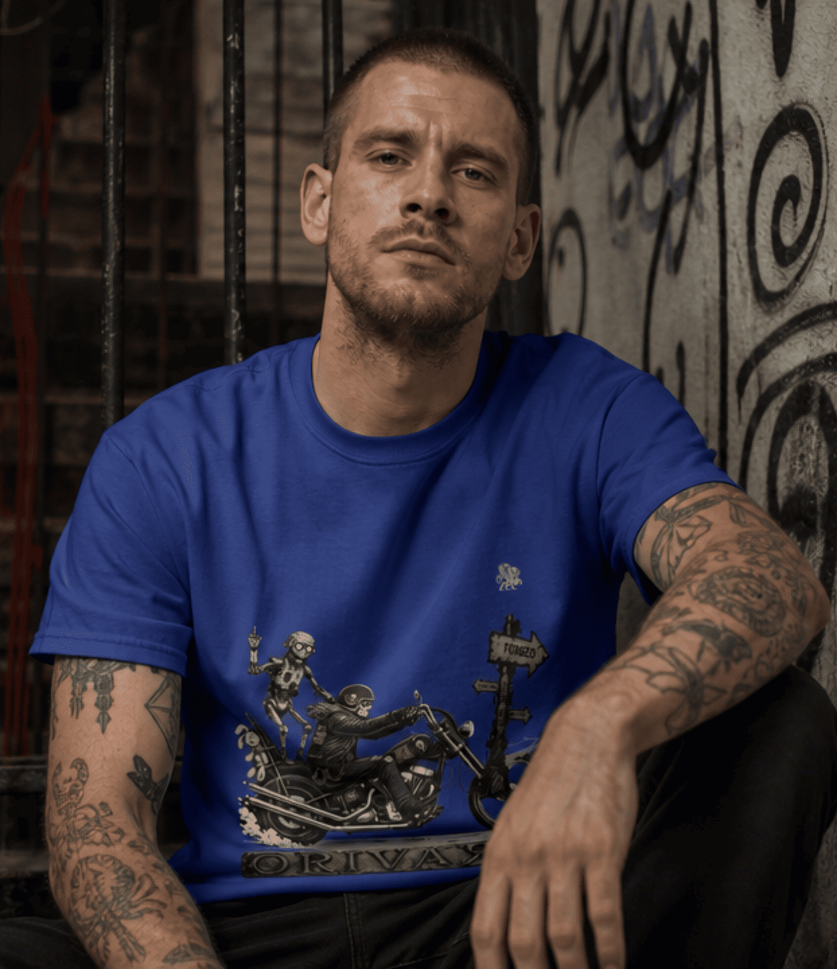 A man with tattoos wearing a royal blue graphic t-shirt featuring a skeleton motorcycle rider design, posed against a dark, graffiti-covered wall.