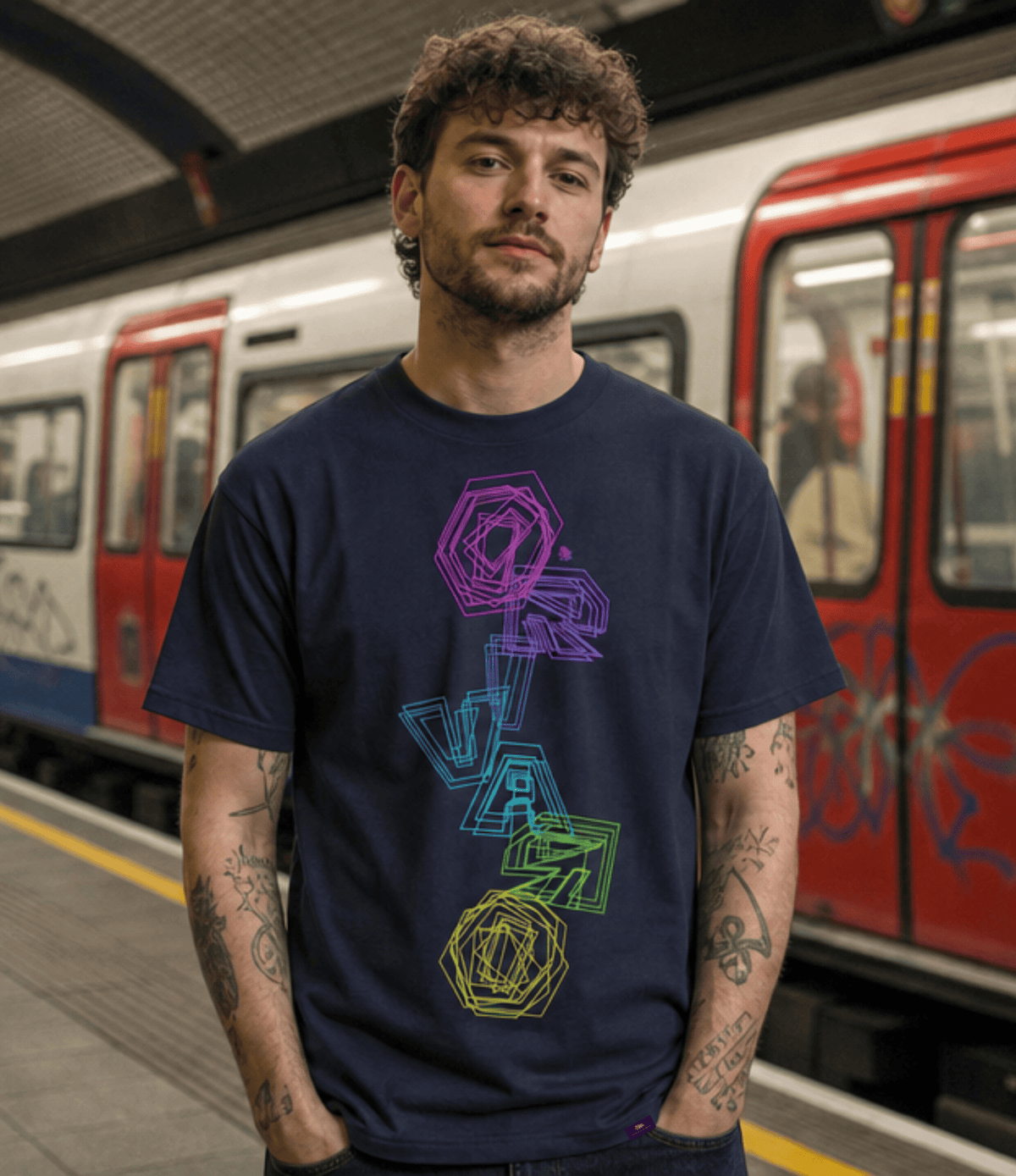 A man with curly brown hair and tattoos stands on a subway platform wearing a navy blue t-shirt with a colourful geometric vertical graphic. A red and white train is blurred in the background.