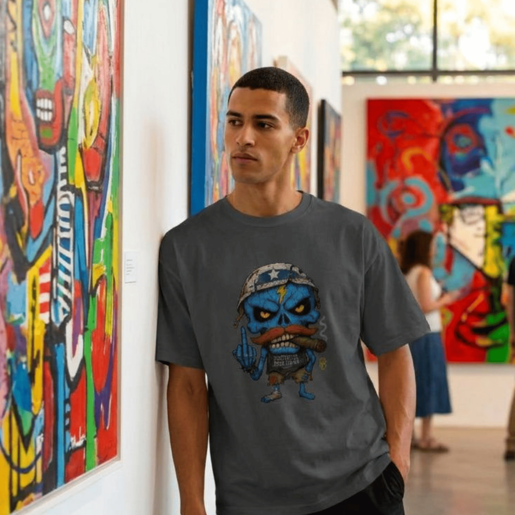 A man wearing a dark grey t-shirt with a cartoon graphic of a blue skull soldier smoking a cigar, standing in an art gallery next to colourful paintings.