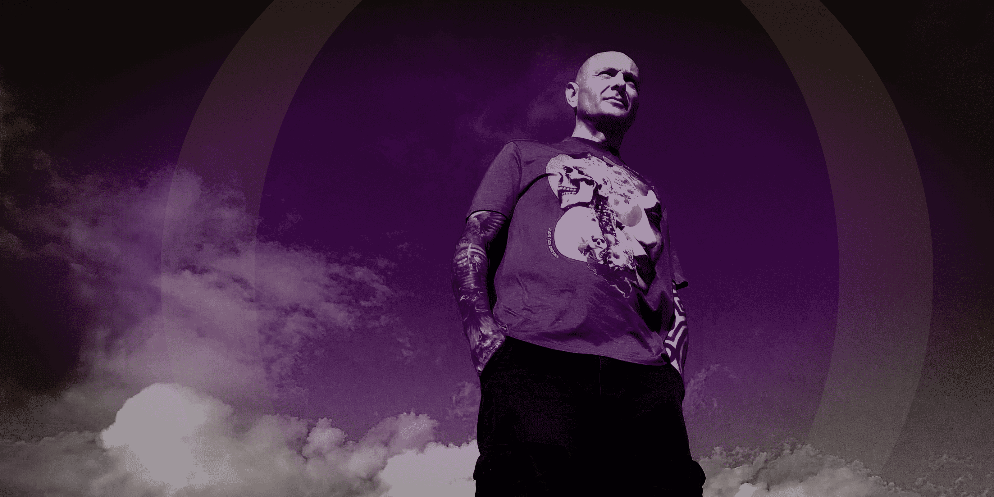 A man wearing a purple graphic t-shirt and black pants, standing against a dark purple background with a circular graphic and clouds.