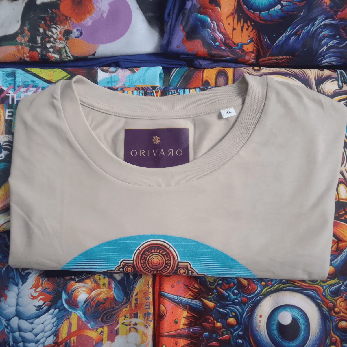 A folded light beige crewneck t-shirt by Orivaro, featuring a graphic print of a stylized eye and turquoise arches across the lower front, laid flat on a highly colourful, patterned background.