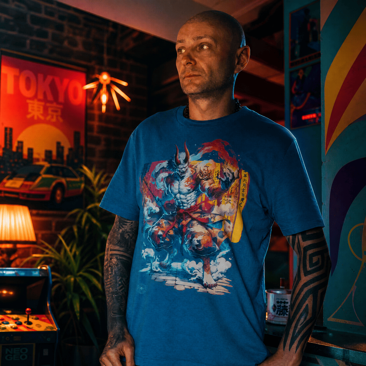 Man wearing a blue t-shirt with a graphic design in a room with neon signs and arcade machines.