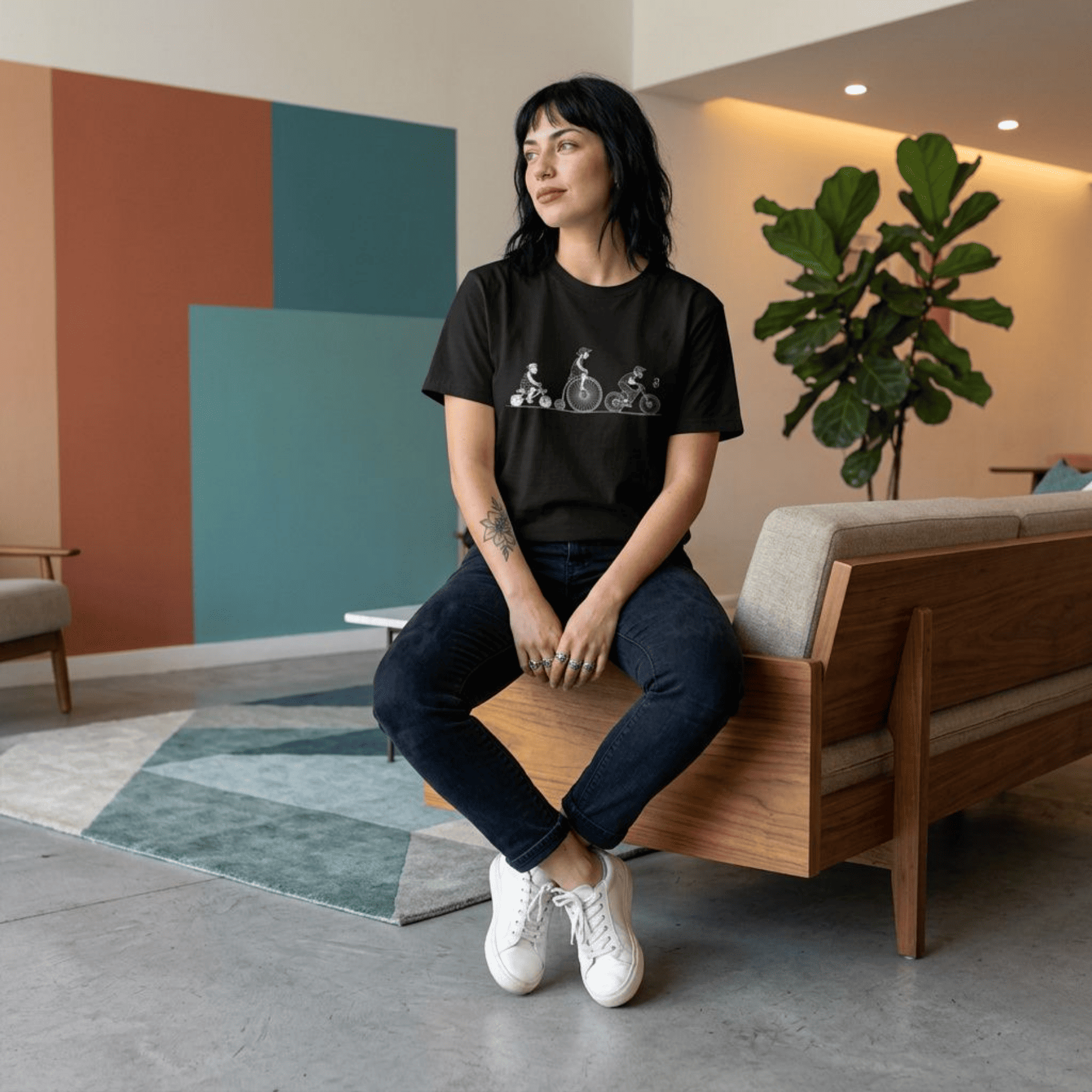 A woman sits on a wooden bench in a modern living room, wearing a black graphic t-shirt, dark jeans, and white sneakers.
