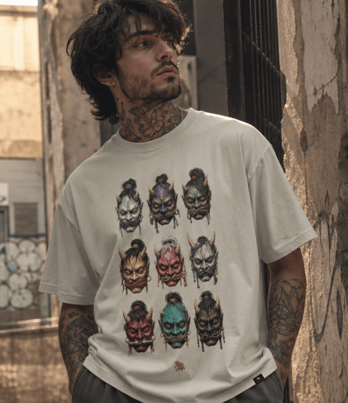 A man wearing a light beige graphic t-shirt with a 3x3 grid of colourful Oni mask prints, standing in an outdoor alleyway.