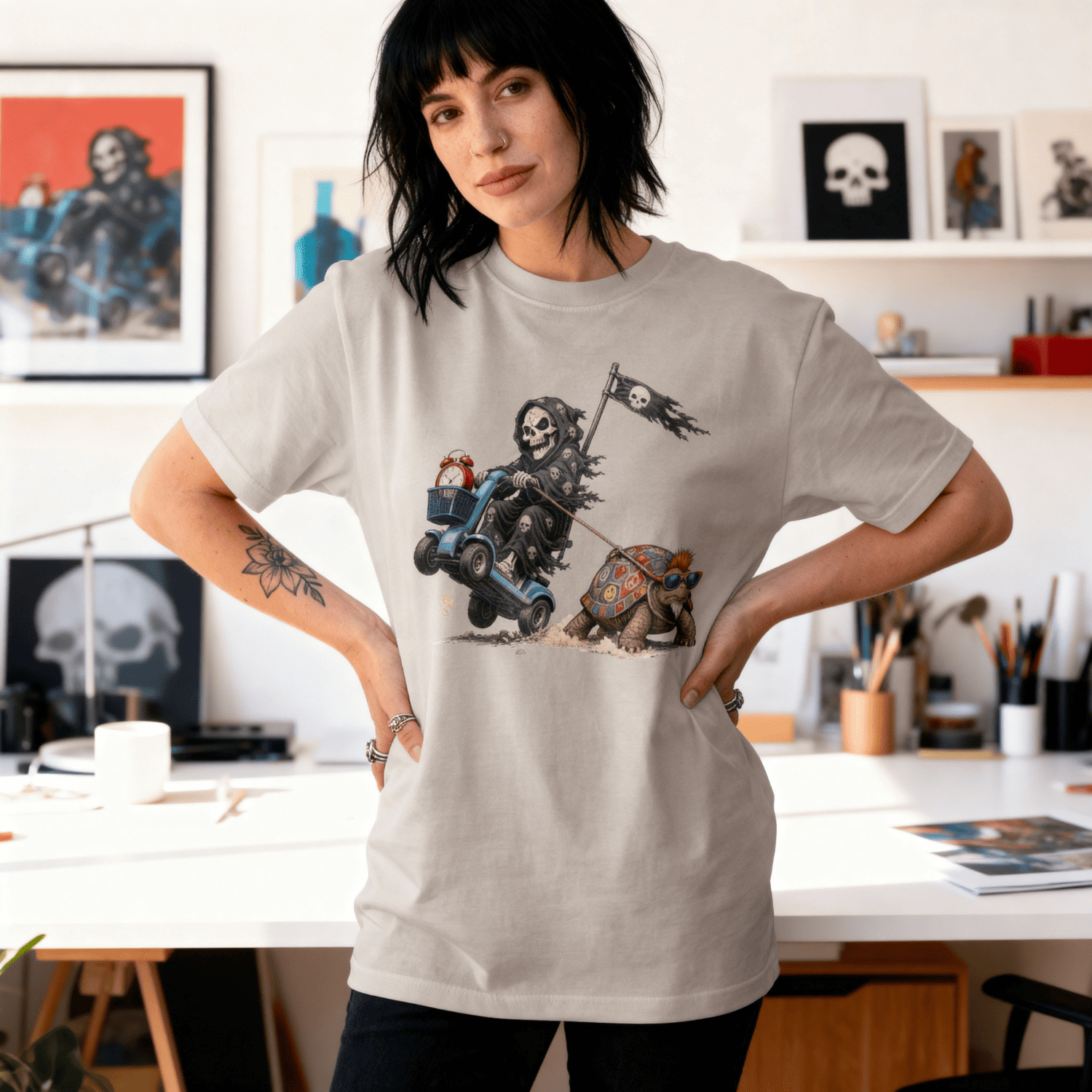 A woman stands in a bright, modern art studio wearing a light grey graphic t-shirt. The t-shirt features a detailed illustration of a skeleton riding a blue motorcycle with a skull flag, accompanied by a small dog.