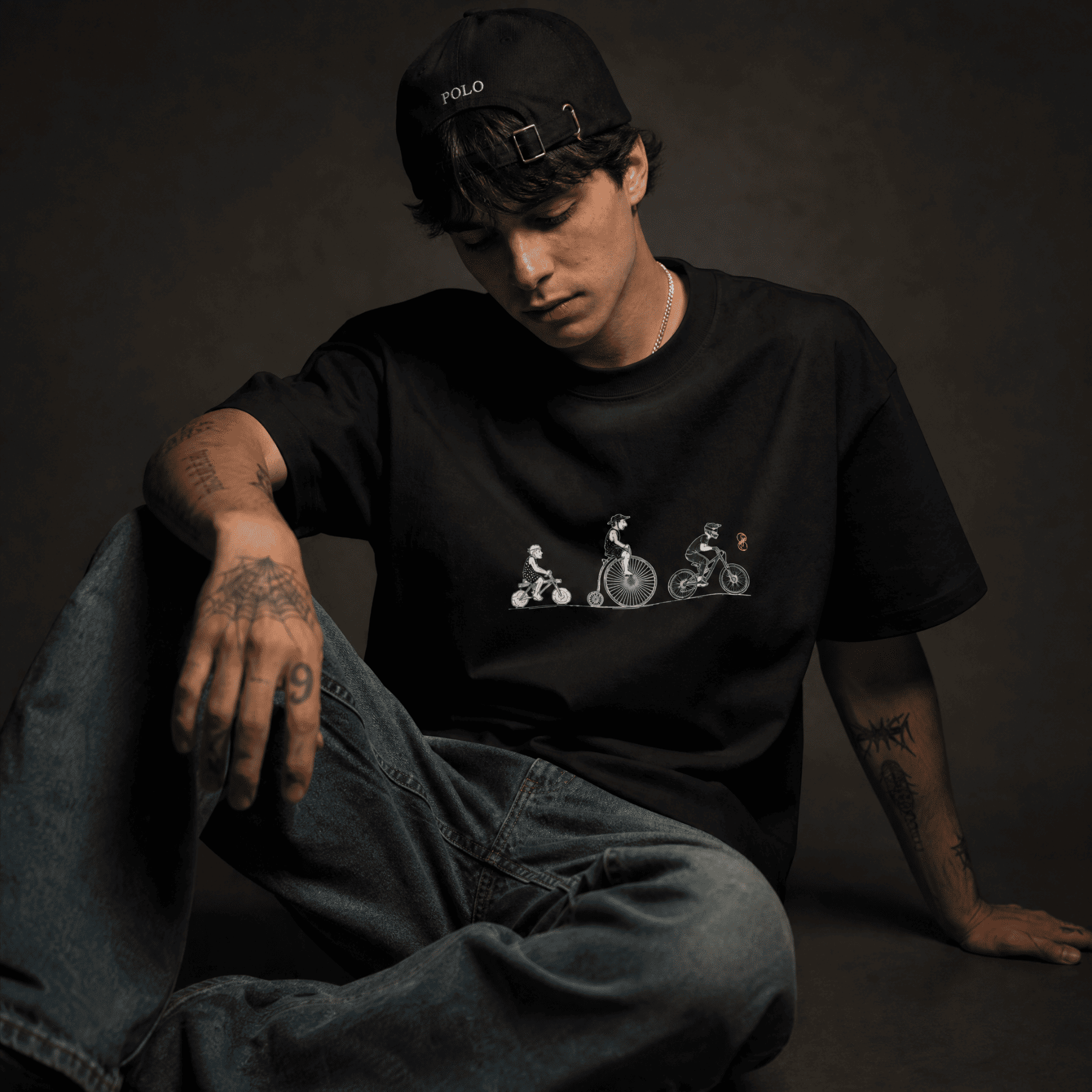 Person wearing a black t-shirt with a graphic design and a cap, sitting against a dark background.
