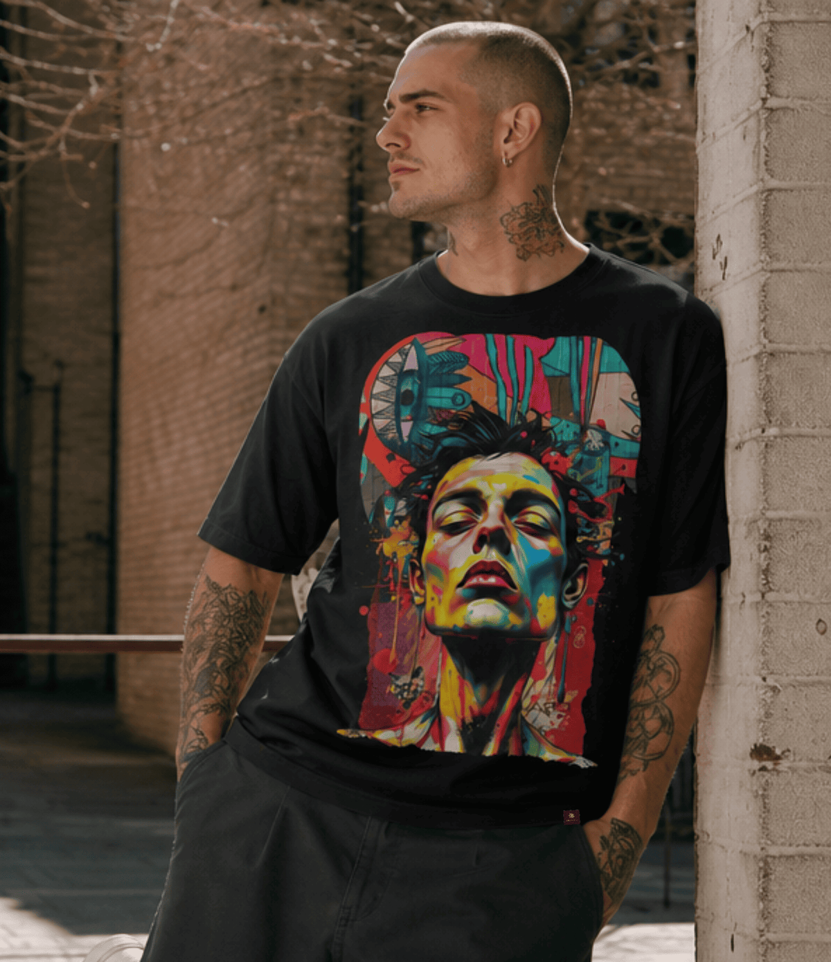 A man wearing a black graphic t-shirt with a colourful, stylized portrait print, leaning against a brick wall in an outdoor urban setting.