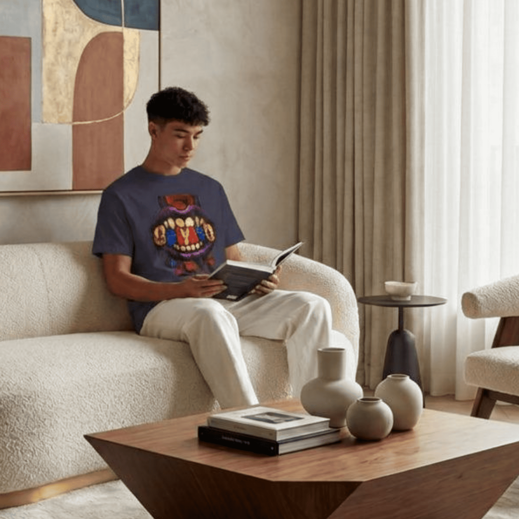 A man sits on a cream boucle sofa in a modern living room, wearing a navy blue graphic t-shirt with a colourful cartoon monster design and white pants, holding an open book.