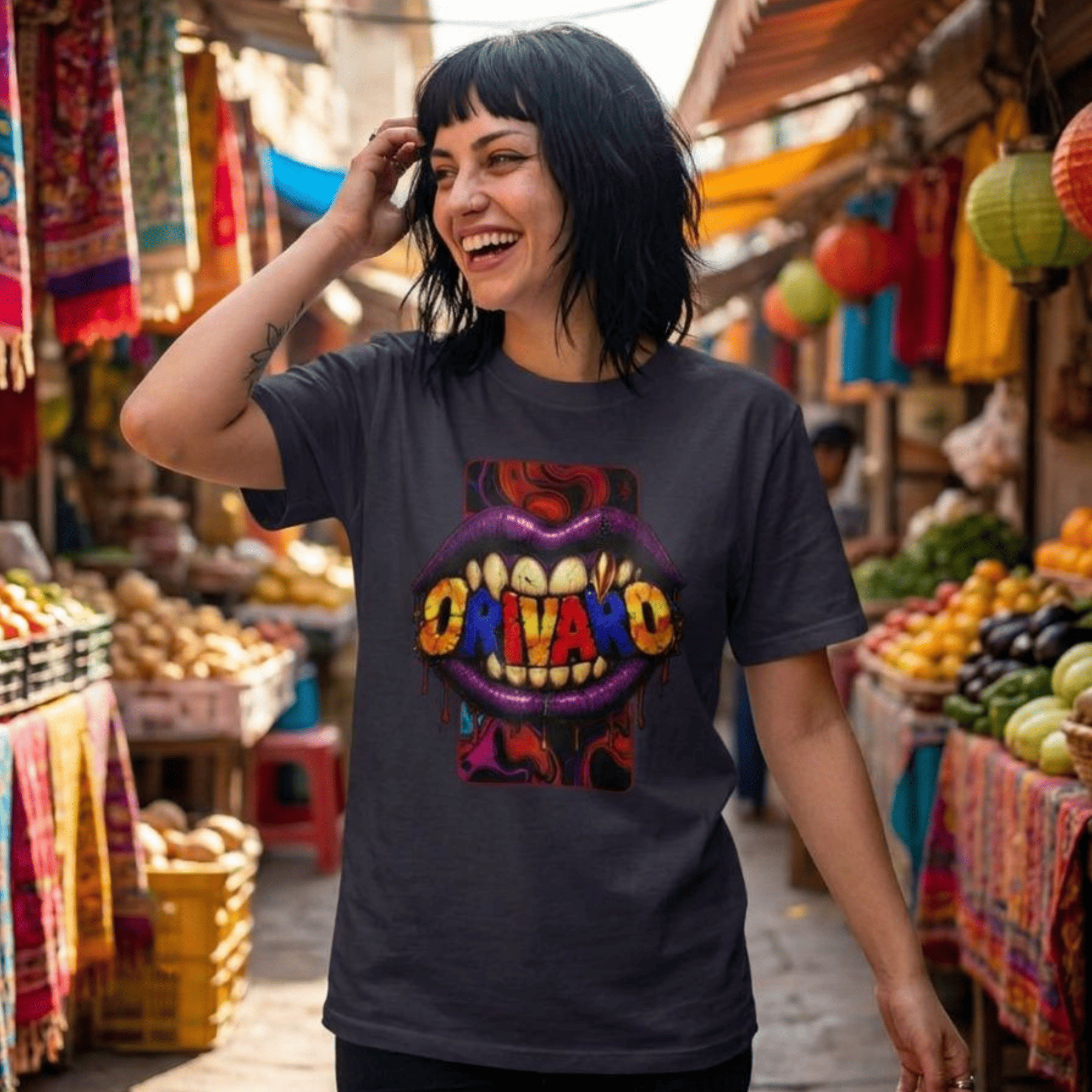 A woman with short black hair stands in a colourful outdoor market wearing a dark charcoal grey graphic t-shirt. The shirt features a vibrant graphic of purple lips with sharp teeth and the word 'ORIVARO' in yellow, red, and blue letters.