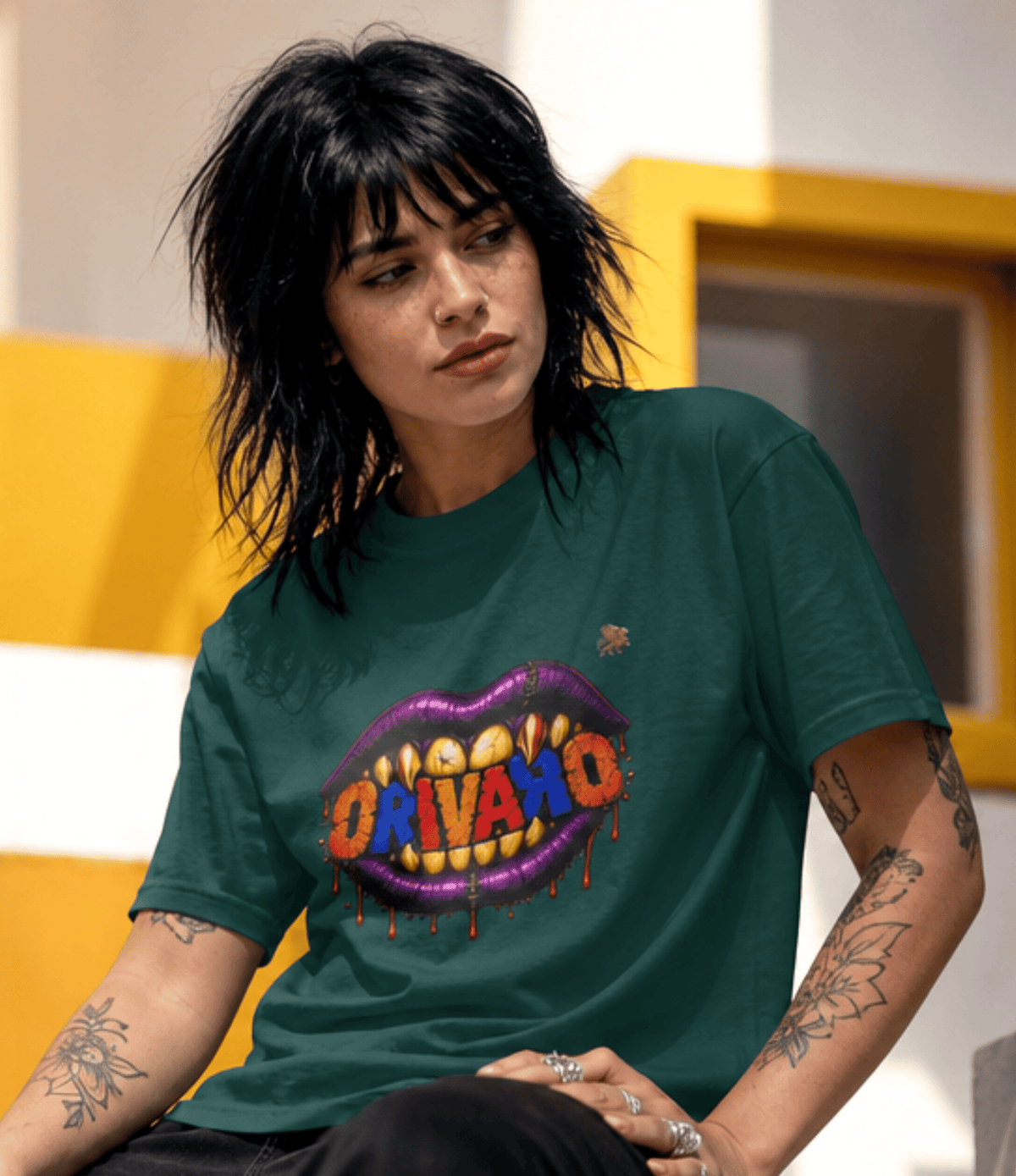 A woman with short dark hair and tattoos wearing a dark green graphic t-shirt. The shirt features a large graphic of purple lips with yellow teeth displaying the word 'ORIVARO' in red and orange letters, with dripping red paint details.