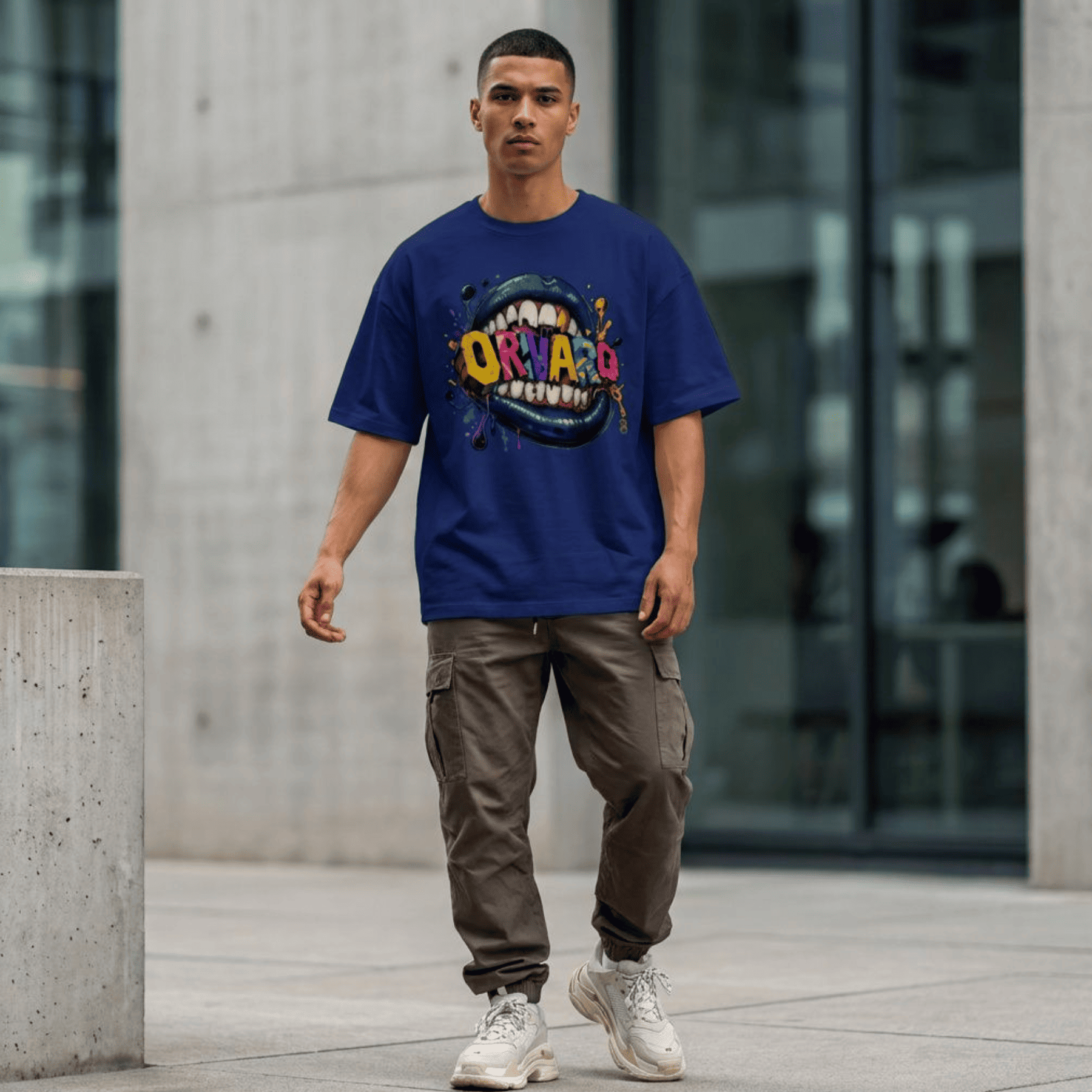 A man walking outdoors in front of a modern concrete building, wearing an oversized royal blue graphic t-shirt with a colourful 'ORIVARO' design, brown cargo pants, and white sneakers.