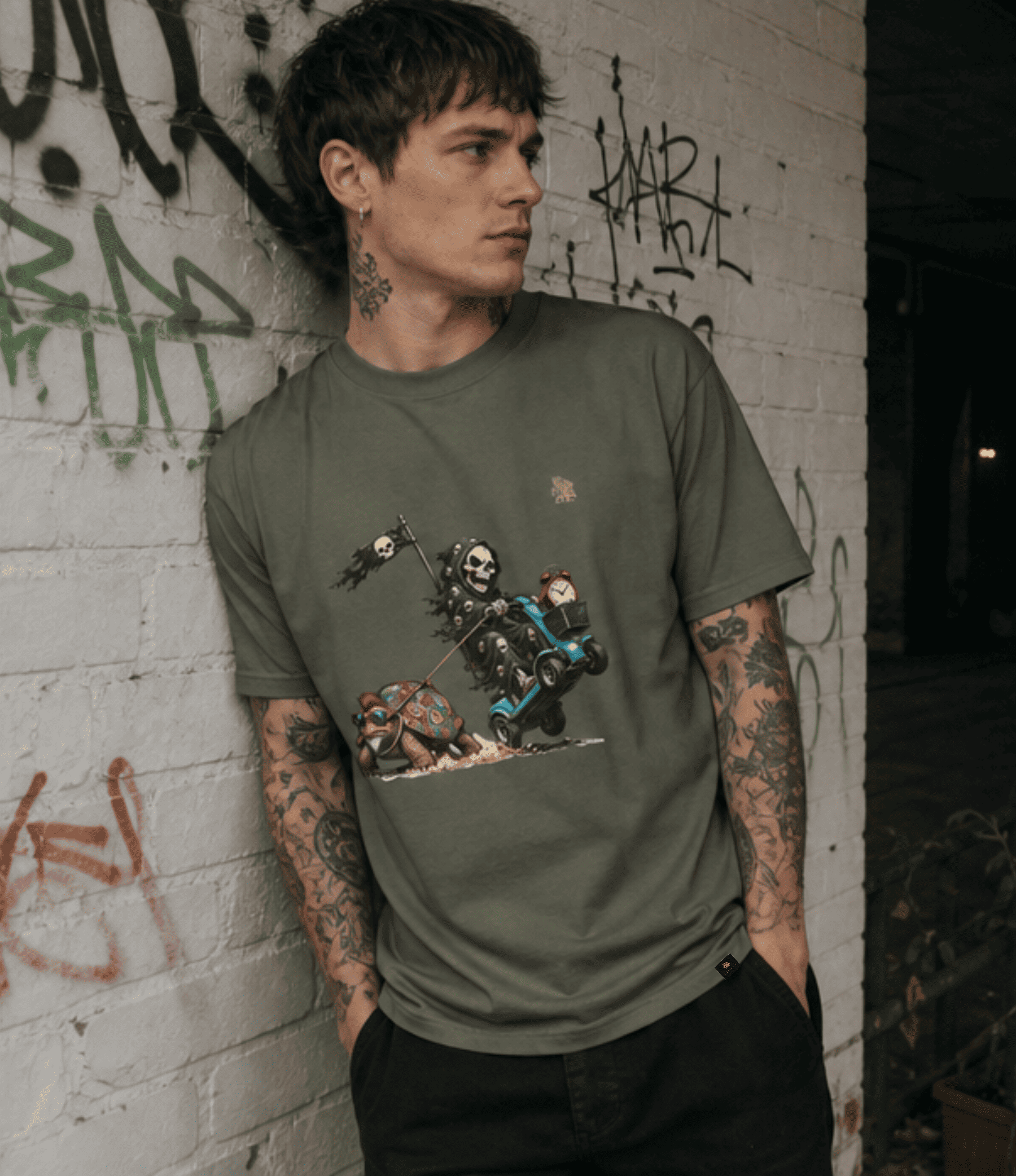 A man with tattoos leans against a graffiti-covered brick wall wearing an olive green graphic t-shirt featuring a cartoon skeleton motorcycle rider.