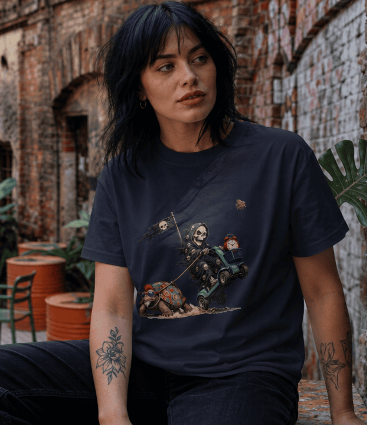 A woman wearing a dark navy graphic t-shirt with a cartoon skeleton and gnomes riding a green cart, posed in front of a rustic brick wall.