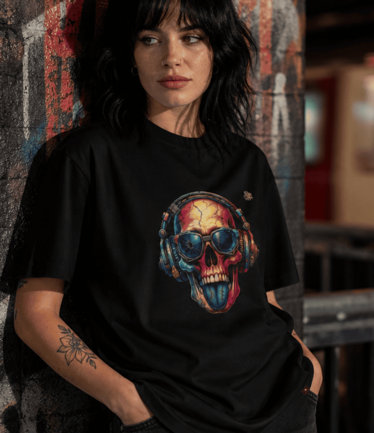 A woman with dark hair leans against a textured wall wearing a black graphic t-shirt. The shirt features a colourful print of a skull wearing headphones and blue aviator sunglasses.