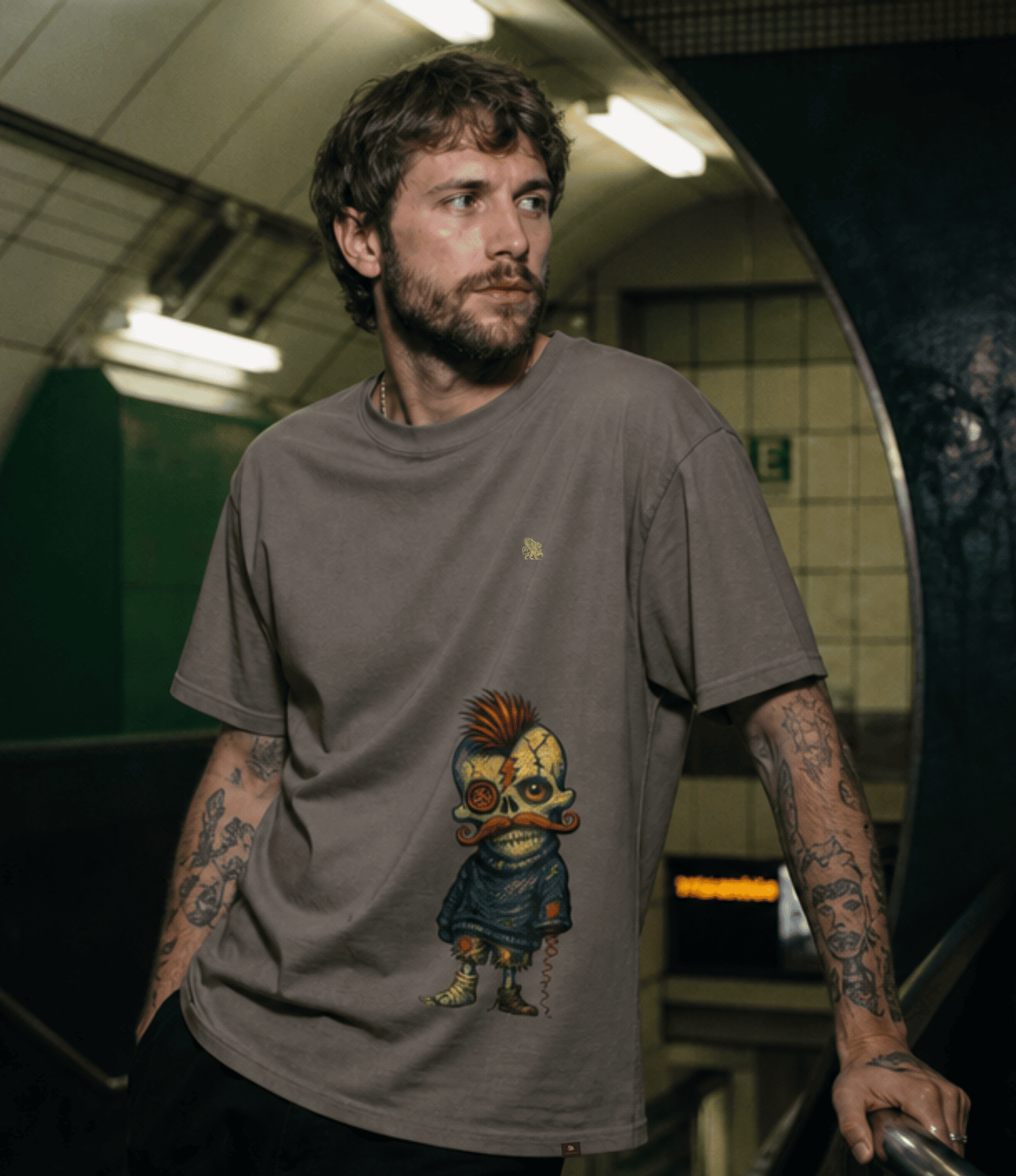 A man with tattoos wearing a brownish-grey graphic t-shirt featuring a cartoon character, standing in a subway station.