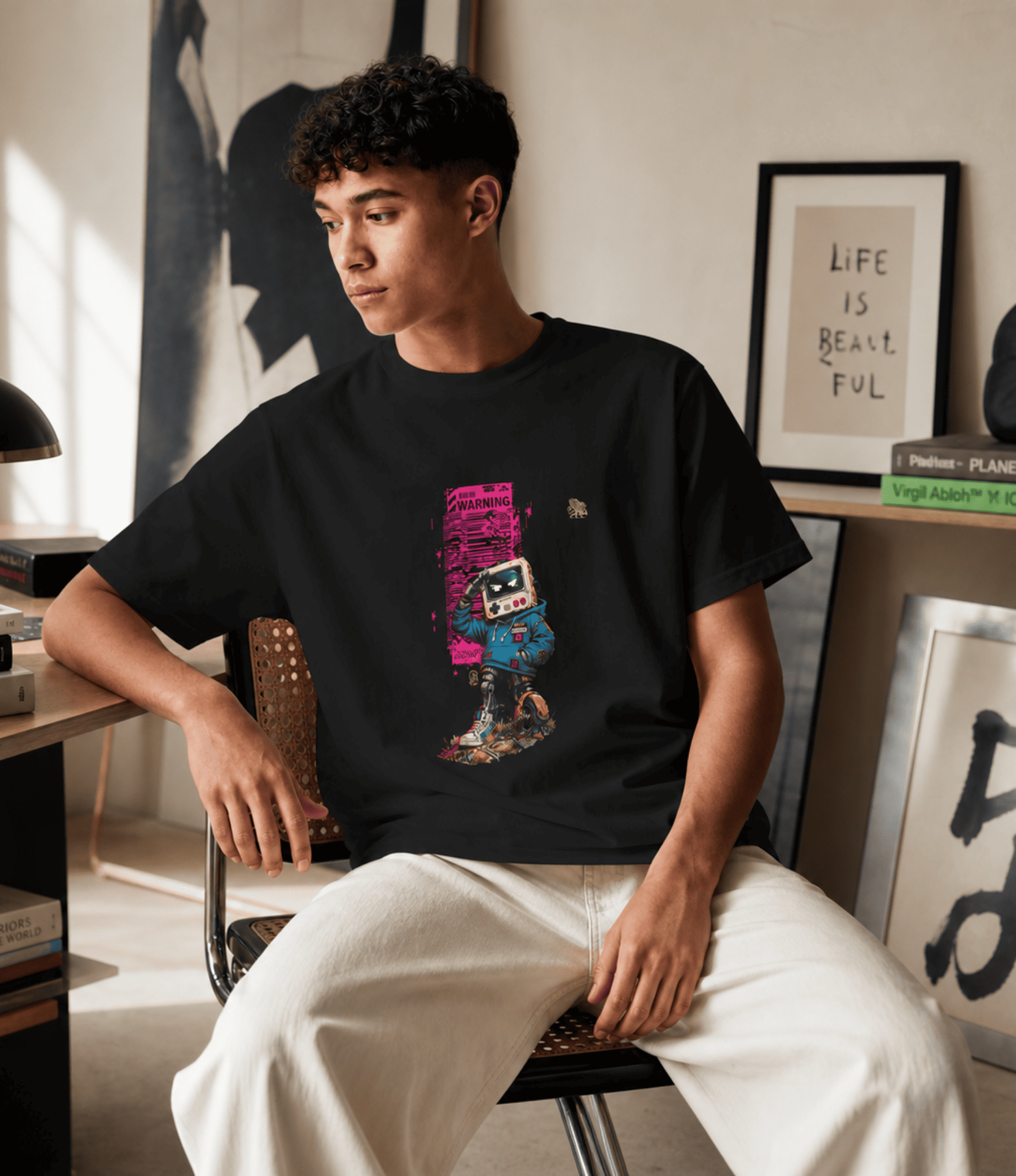 A man sitting in a chair wearing a black graphic t-shirt with a colourful retro cassette-tape design and white pants, in a modern, sunlit room with framed art.