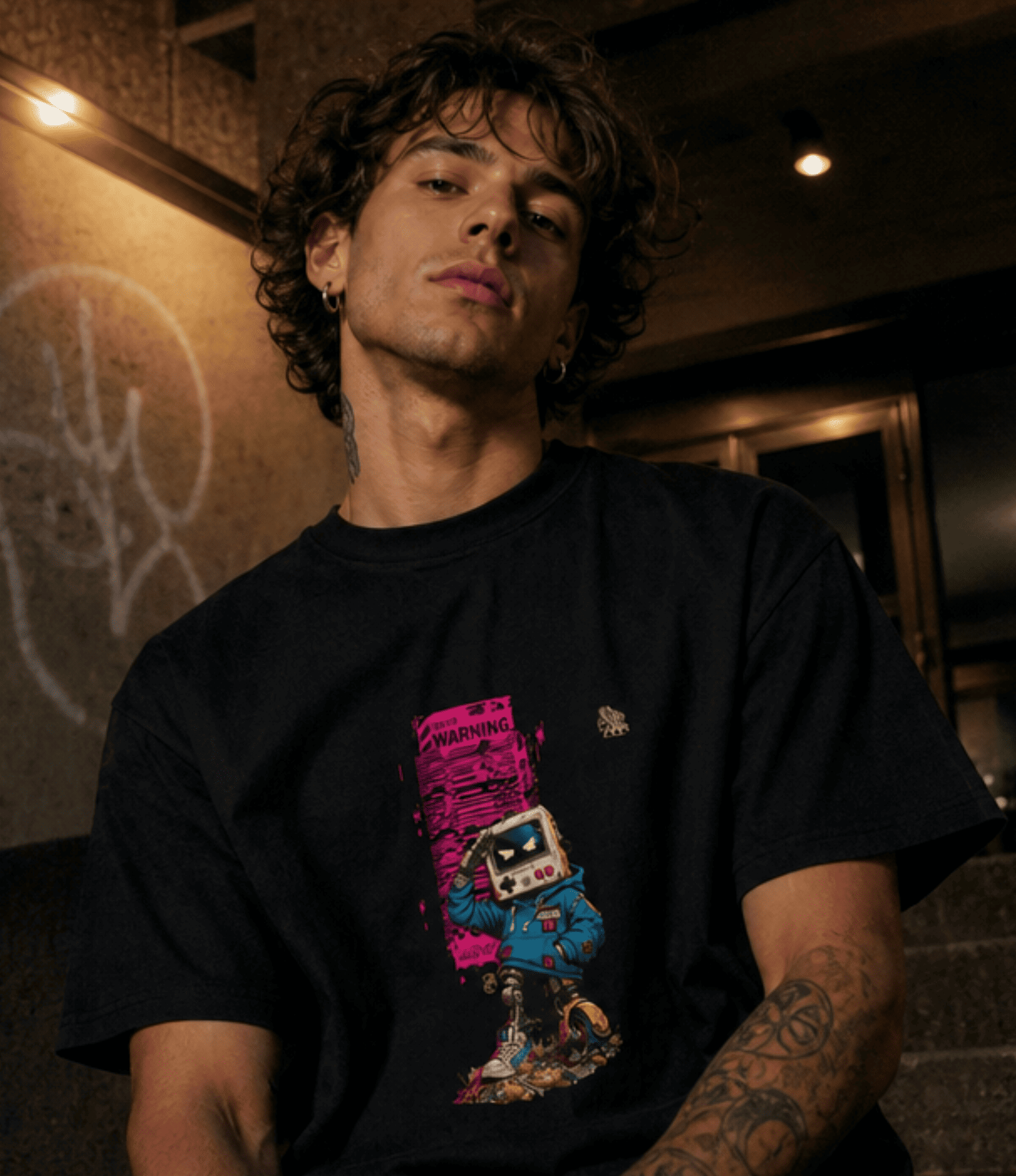 A man wearing a black graphic t-shirt with a colourful robot design, photographed in a dimly lit indoor environment.