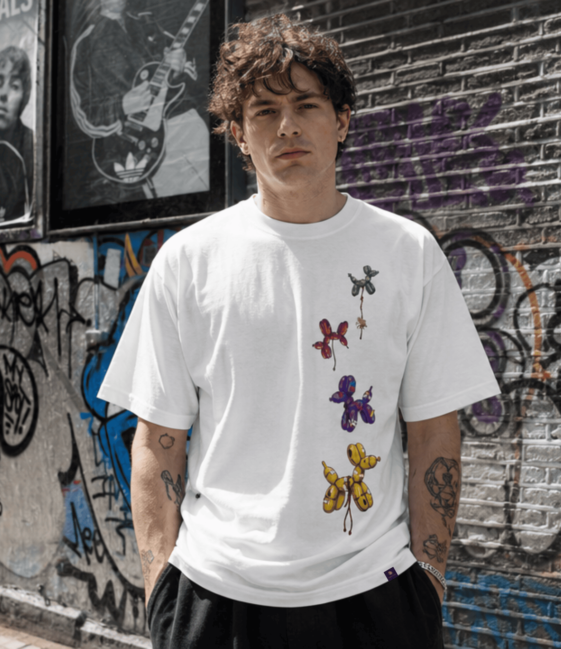 A man wearing a white crewneck t-shirt with a vertical graphic print of colourful balloon dogs, standing in front of a graffiti-covered brick wall.