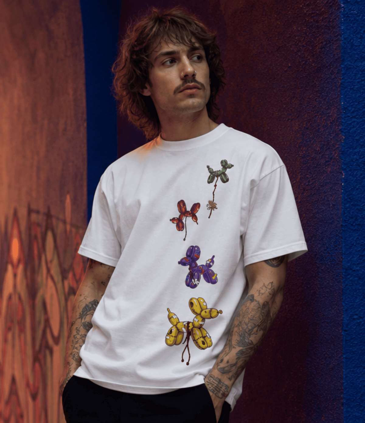 A man with curly brown hair and tattoos wearing a white t-shirt with a vertical graphic of balloon-dog illustrations in green, red, purple, and yellow, posed against a textured wall with graffiti and blue and orange panels.