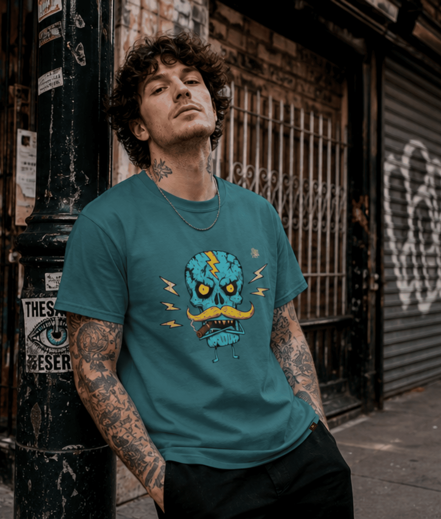 A man with curly hair and tattoos leans against a distressed black pole on a city street, wearing a teal graphic t-shirt with a cartoon skull wearing a mustache.