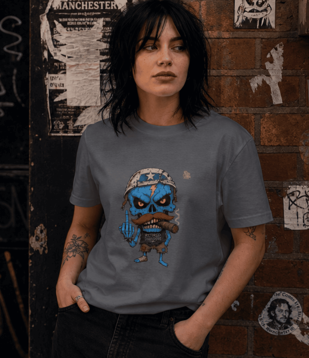 A woman with short dark hair stands against a brick wall with posters, wearing a grey graphic t-shirt. The t-shirt features a distressed blue cartoon skull character wearing a military helmet and holding a cigar.