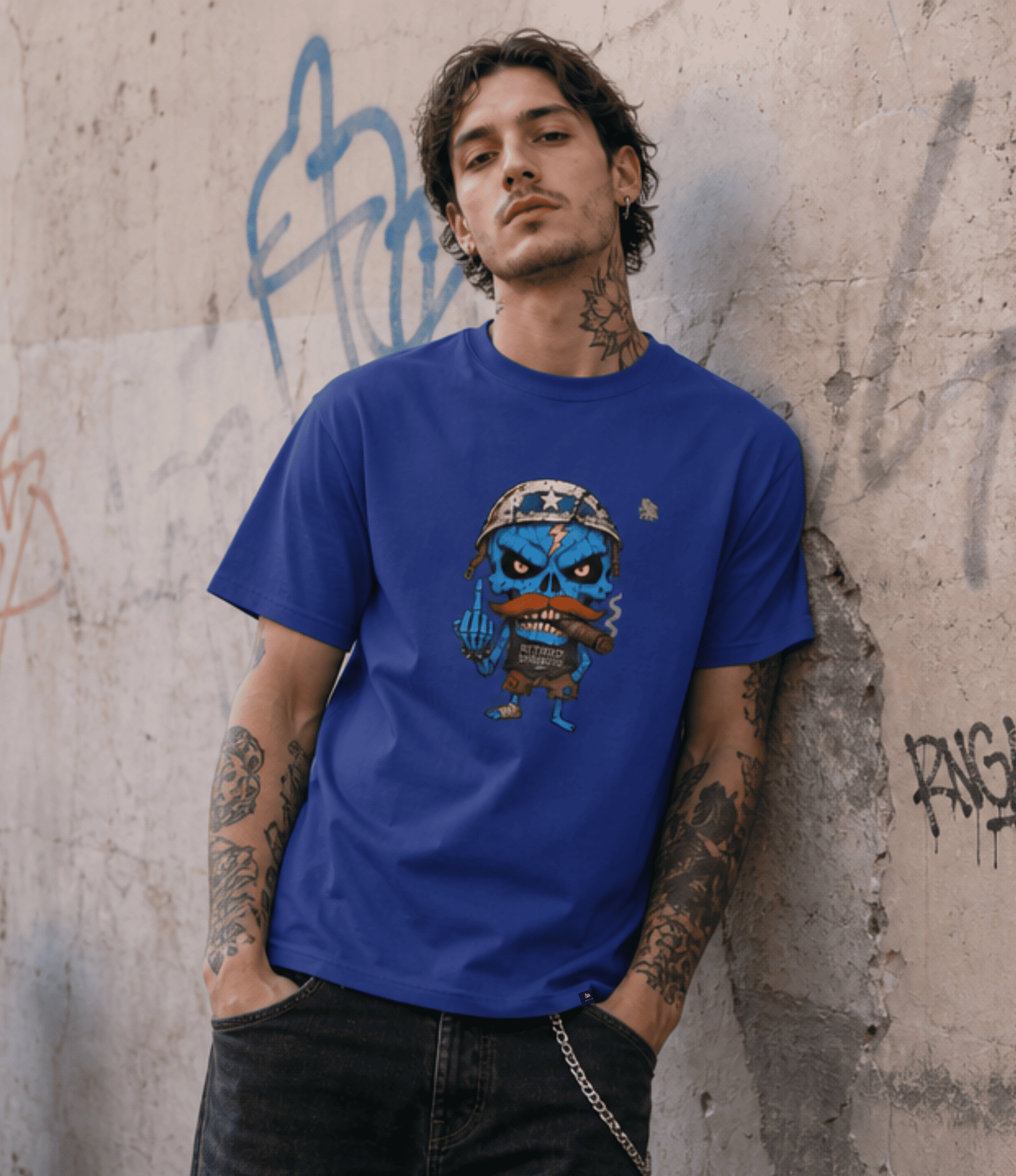 A man with tattoos leans against a concrete wall with graffiti, wearing a royal blue crewneck t-shirt featuring a cartoon graphic of a blue skull character with a mustache.