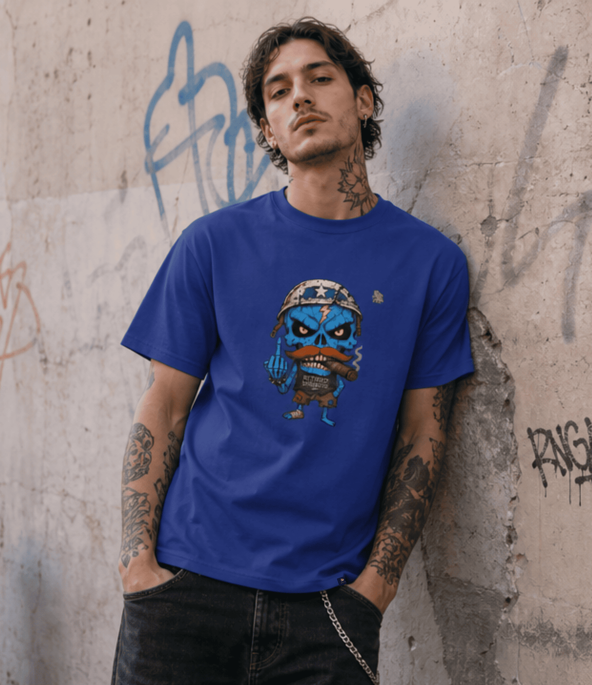 A man with tattoos leans against a concrete wall with graffiti, wearing a royal blue crewneck t-shirt featuring a cartoon graphic of a blue skull character with a mustache.