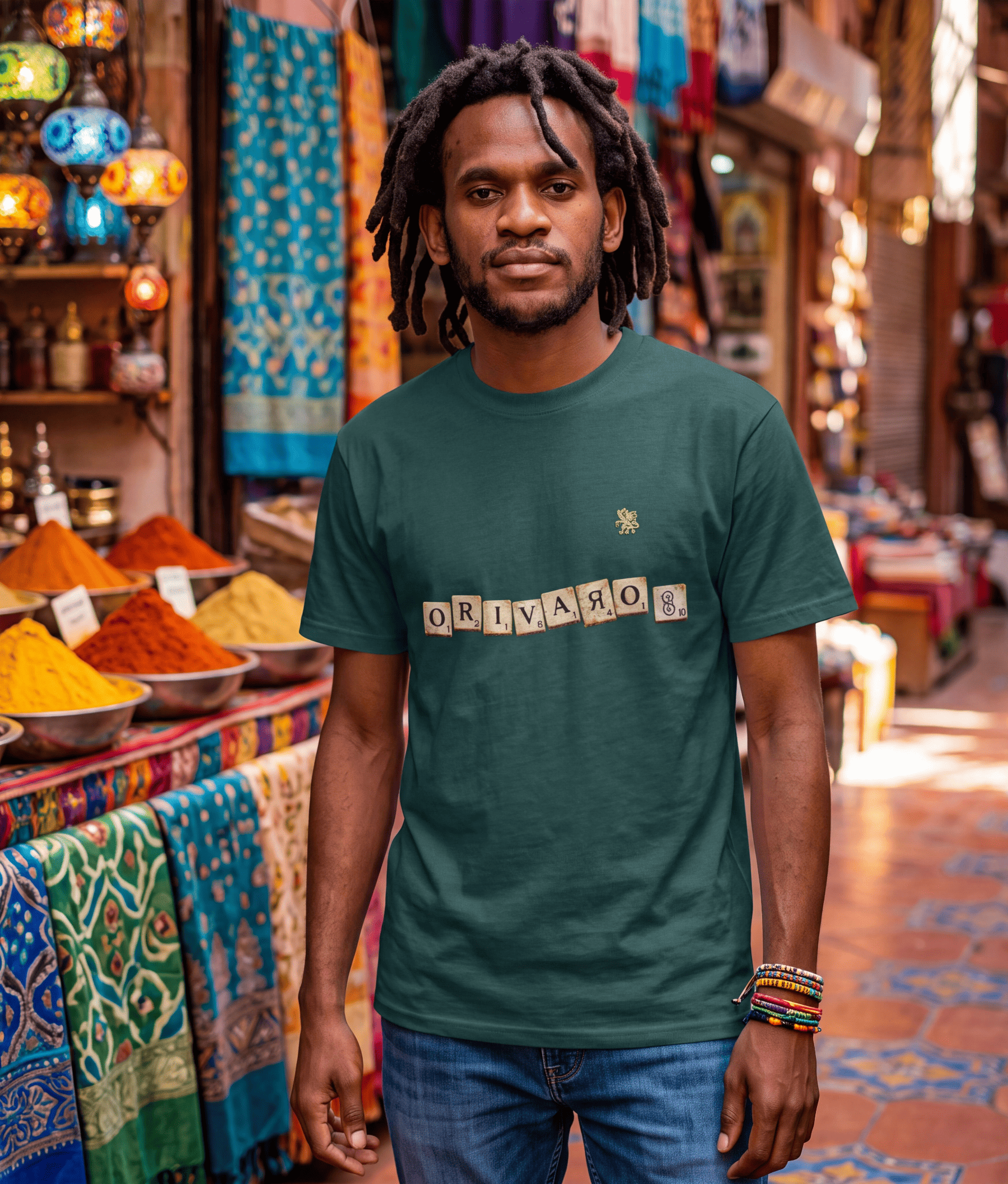 A man with dreadlocks sits in a wooden armchair in a cozy, book-filled room, wearing a dark teal t-shirt with the word 'ORIVARO' spelled out in wooden toy blocks on the chest.