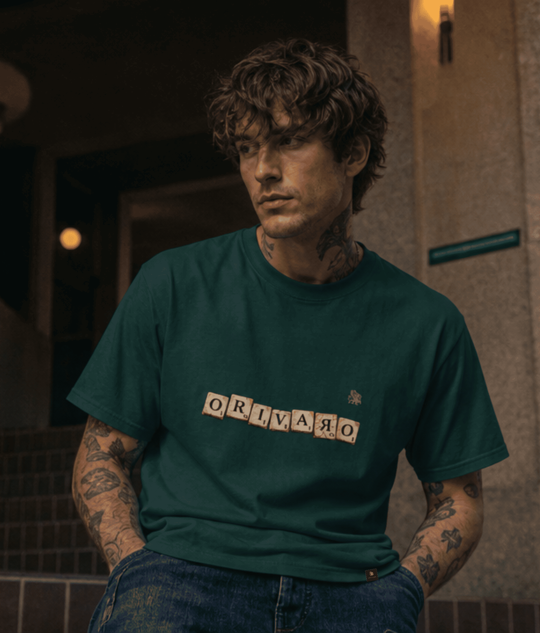 A man wearing a dark green crewneck t-shirt with 'ORIVARO' spelled out in block letters across the chest, paired with blue jeans in a dimly lit indoor setting.