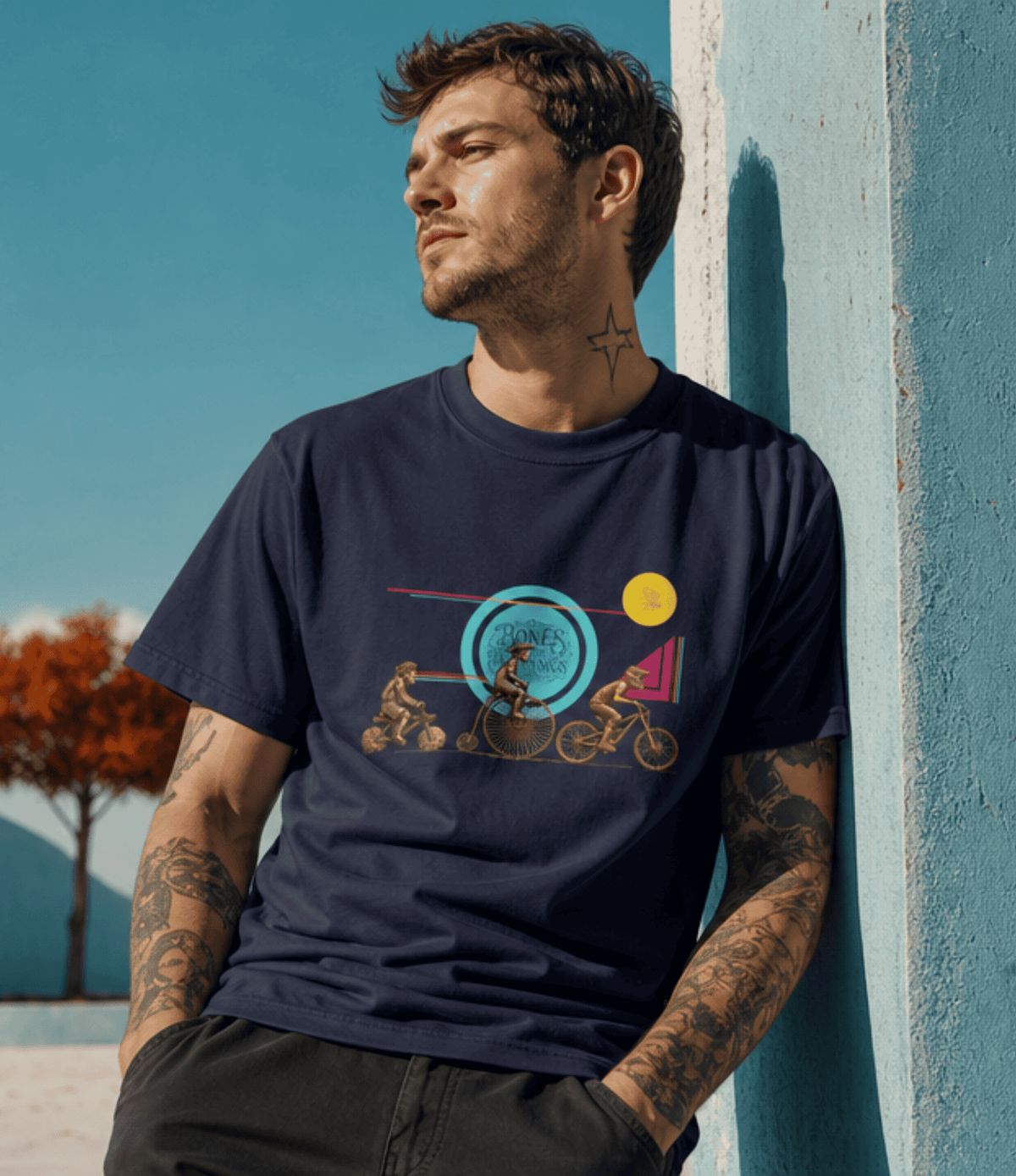 A man with tattoos leans against a textured blue wall, wearing a navy blue graphic t-shirt with a retro illustration of two figures and geometric shapes, set against a sunny outdoor background.