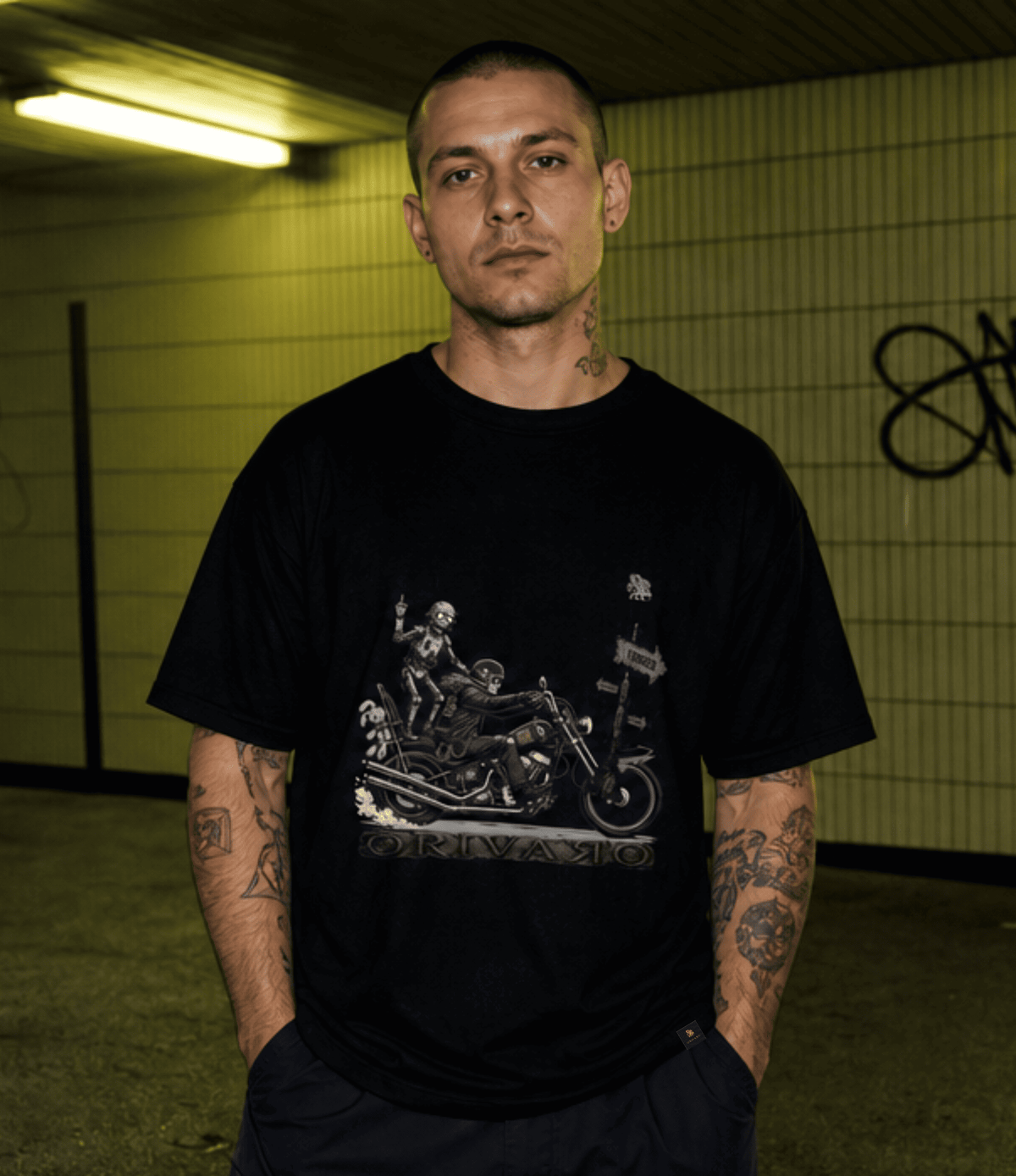 A man with tattoos wearing a black graphic t-shirt featuring a skeleton riding a chopper motorcycle and the word 'ORIVARO' printed below it, standing in a tiled indoor space - tube station.