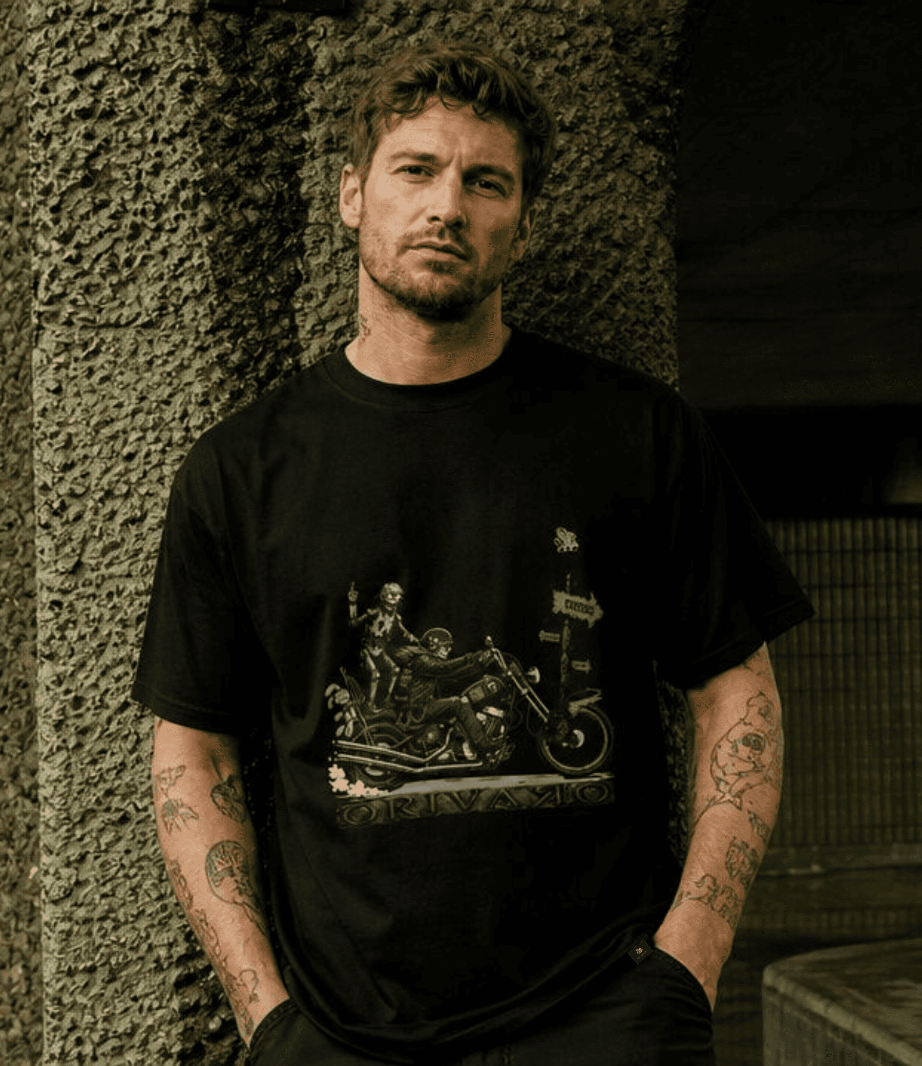 A man with tattoos wearing a black graphic t-shirt with a motorcycle and skeleton design, standing against a textured concrete wall.