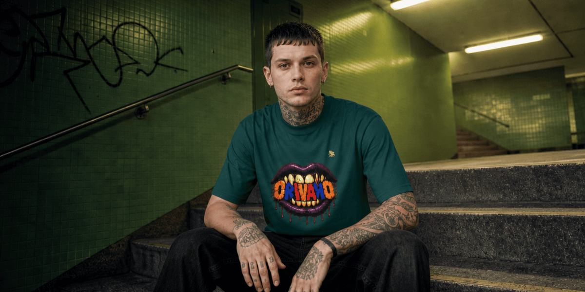 A tattooed man sitting on concrete stairs in a green tiled indoor space, wearing a dark green t-shirt with a colourful graphic of a mouth with teeth spelling 'ORIVARO.