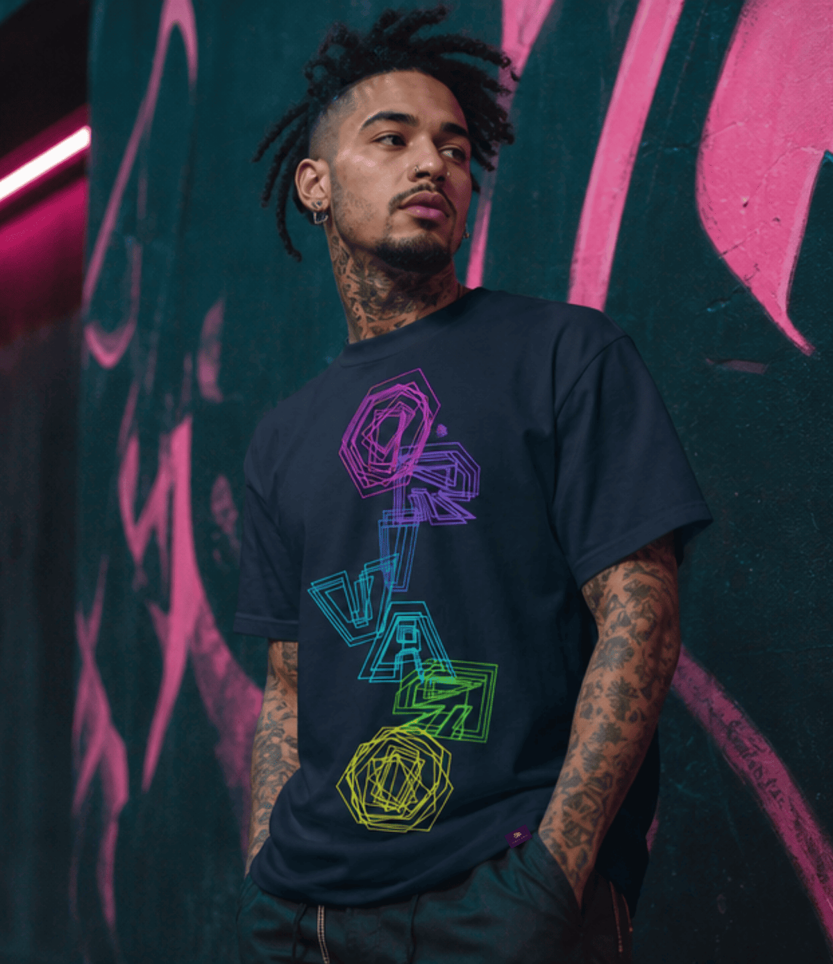 A man with tattoos and dreadlocks wearing a black t-shirt with a colourful geometric 'ORIVARO' graphic, standing against a dark wall with pink graffiti.
