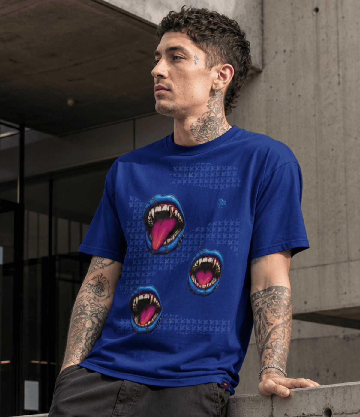 A man wearing a royal blue graphic t-shirt with three shark mouth prints, leaning against a concrete wall in an urban setting.