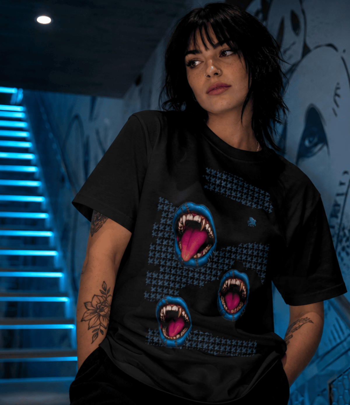A man wearing a navy blue graphic t-shirt with three blue and pink screaming mouth designs, posed against a yellow graffiti-covered wall.