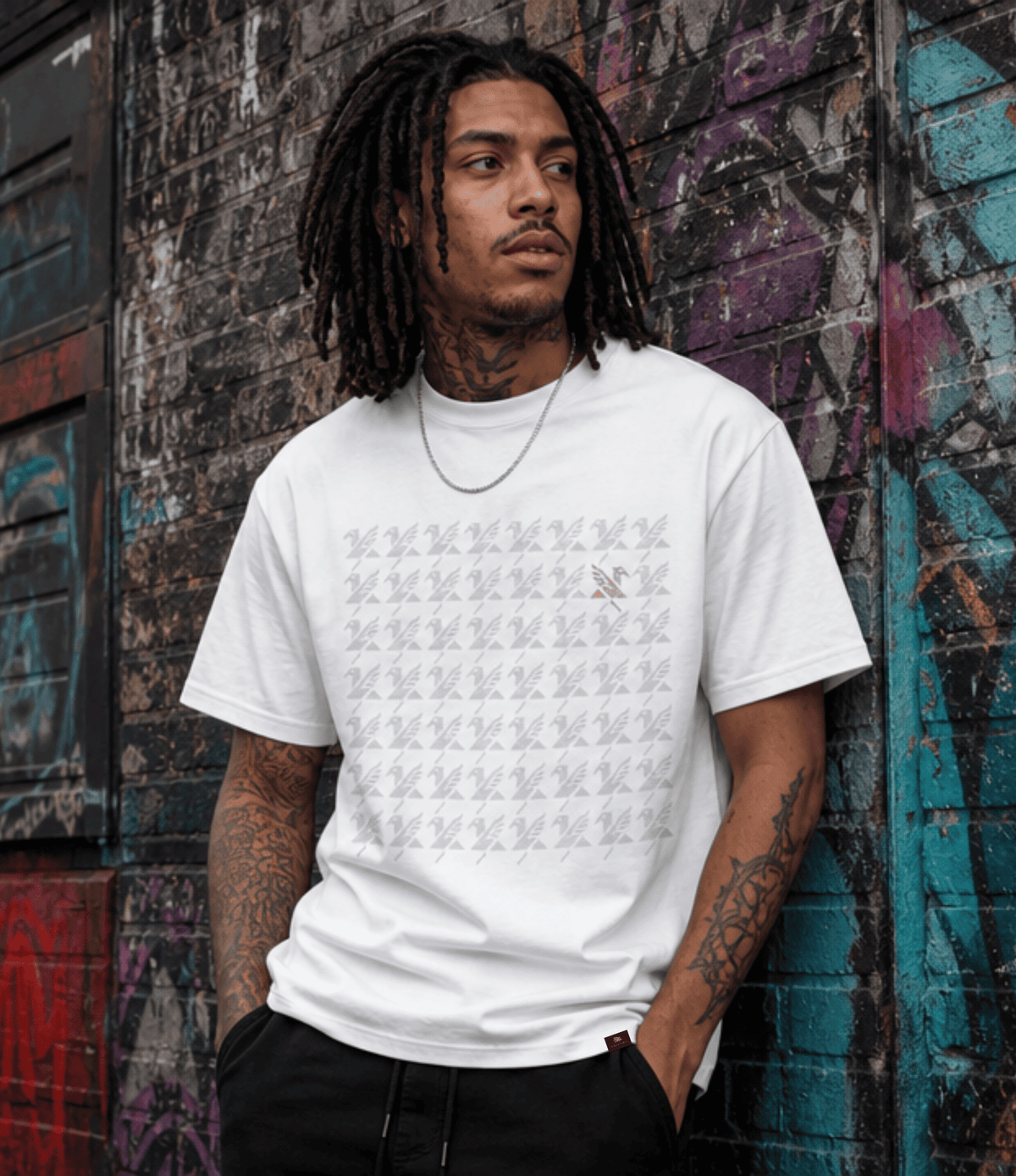 A man with dreadlocks stands against a graffiti-covered brick wall wearing a white graphic t-shirt with a grid pattern and a small griffin logo on the chest, paired with black pants.