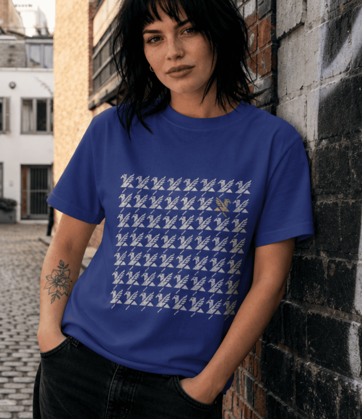 A woman wearing a blue crewneck t-shirt with a white graphic print of repeated 'griffin' shapes, leaning against a brick wall in an outdoor urban setting.