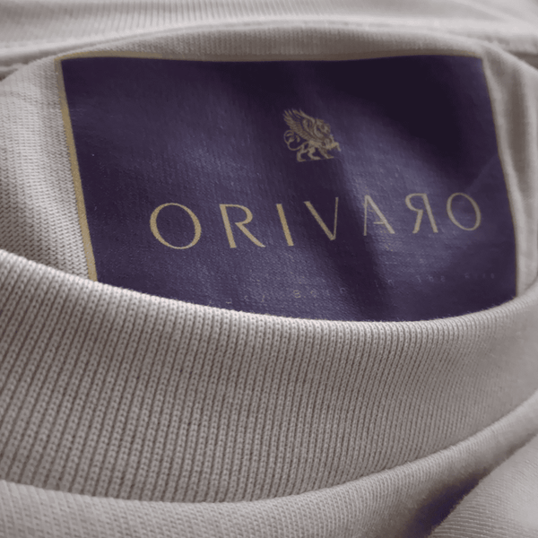 Close-up of a light beige ribbed knit garment showing the interior neck label with a dark purple background and gold lettering reading ORIVARO.