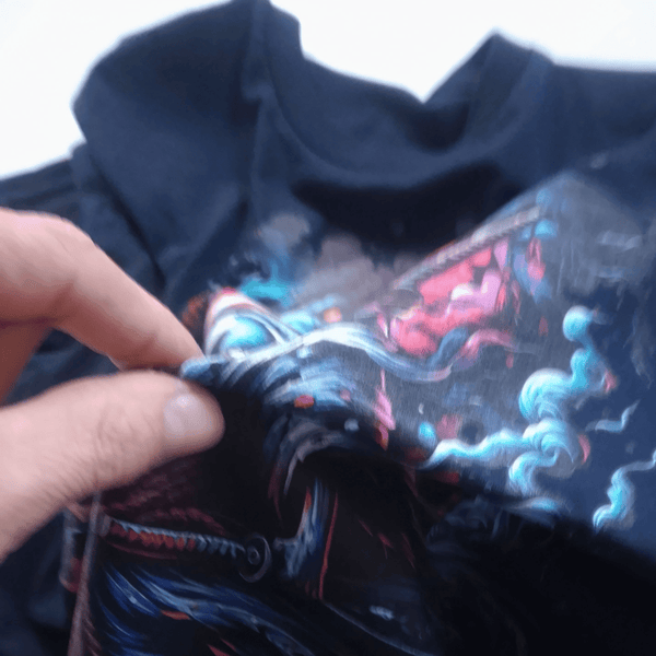A close-up of a black t-shirt being held by a hand, showing a detailed graphic print with swirling blue, red, and white smoke-like patterns.