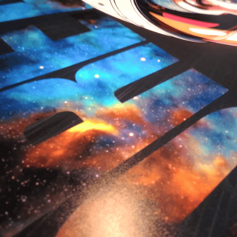 A close-up of a printed surface featuring a colourful galaxy or nebula design with blue, orange, black, and white star-like areas, partially overlaid by dark piano-key-like shapes and a blurred reflective circular object at the top.